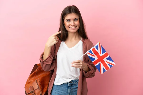 年轻的高加索女人举着一面英国国旗 背景是粉红色的 她举起大拇指做了一个手势 — 图库照片