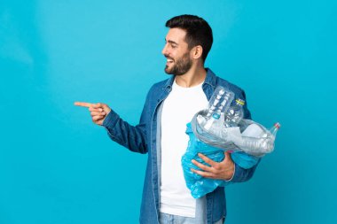 Mavi arka planda izole edilmiş plastik şişelerle dolu bir çanta taşıyan genç beyaz bir adam bir ürünü sunmak için kenarı işaret ediyor.
