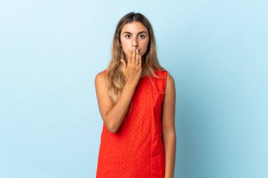 İzole edilmiş mavi arka planda, ağzı eliyle kapatan genç İspanyol kadın.