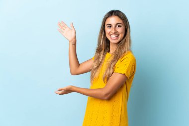 İzole edilmiş mavi arka planda genç bir İspanyol kadın davet etmek için ellerini yana uzatıyor.