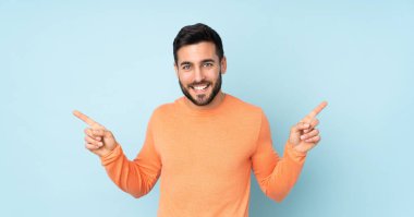 Beyaz tenli yakışıklı adam parmak uçlarını işaret ediyor ve mavi arka planda mutlu.