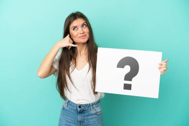 Kafkasyalı genç bir kadın mavi arka planda izole edilmiş. Elinde üzerinde soru işareti olan bir levha var ve telefon hareketi yapıyor.