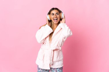 Pijamalı, pembe arka planda, elinde yastıkla müzik dinleyen genç bir İspanyol kadın.