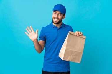Mavi arka planda sakallı teslimatçı. Mutlu bir ifadeyle selamlıyor.