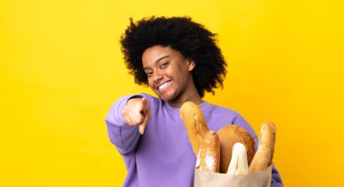 Genç Afrikalı Amerikalı bir kadın sarı arka planda izole edilmiş bir şey alıyor. Önü mutlu bir ifadeyle işaret ediyor.