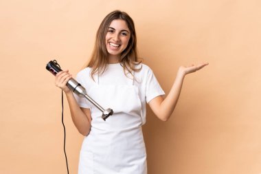 İzole edilmiş arka planda el blenderi kullanan kadın davet edilmek için ellerini yana açıyor.