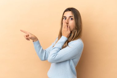Beyazlı genç bir kadın bej arka planda izole edilmiş, sürpriz bir ifadeyle işaret ederken...