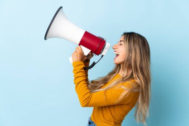 İzole edilmiş mavi arka planda genç İspanyol kadın megafondan bir şey duyurmak için bağırıyor.