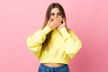 Soyutlanmış pembe arka planı olan genç bir kadın elleriyle ağzını kapatıyor.