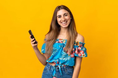 İzole edilmiş sarı arka planda cep telefonu kullanan genç bir kadın çok fazla gülümsüyor.