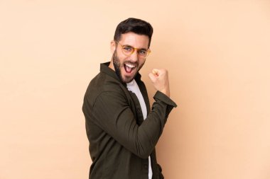 Beyaz tenli yakışıklı adam bej arka planda izole edilmiş bir şekilde zaferi kutluyor.