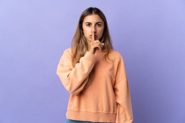 İzole edilmiş mor arka planda genç İspanyol kadın parmağını ağzına koyan sessizlik işareti gösteriyor.