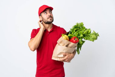 Sakallı genç bir adam elinde bir çanta dolusu sebzeyle beyaz arka planda izole edilmiş bir fikir düşünüyor.