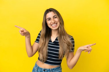 Sarı arka planda izole edilmiş genç beyaz kadın parmak uçlarını işaret ediyor ve mutlu.