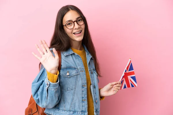 Young French girl holding an United Kingdom flag isolated on pink background saluting with hand with happy expression