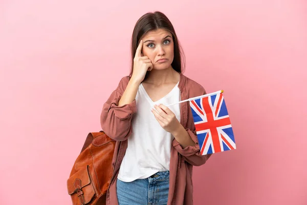 一个年轻的高加索女人举着一面英国国旗 与粉色背景隔离 思考着一个想法 — 图库照片