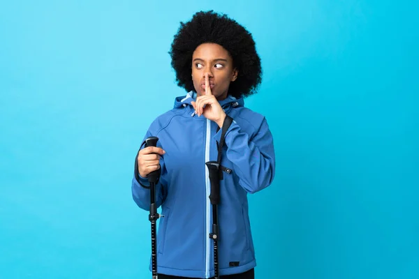 若いアフリカ系アメリカ人のバックパックと青の背景に隔離されたトレッキングポールが口の中に指を入れて沈黙ジェスチャーの兆候を示しています — ストック写真
