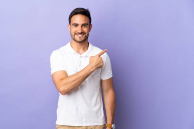 Mor arka planda izole edilmiş genç yakışıklı bir adam bir ürün sunmak için kenarı işaret ediyor.