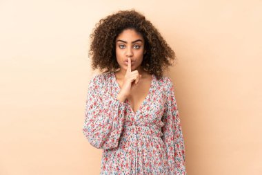 Genç Afrikalı Amerikalı kadın bej arka planda izole edilmiş. Sessiz bir jest işareti var. Parmağını ağzına sokuyor.