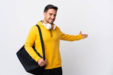 Spor çantalı genç adam beyaz arka planda izole edilmiş. Davet etmek için ellerini yana uzatıyor.