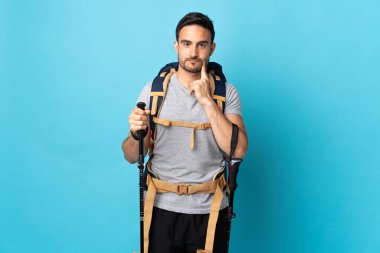 Sırt çantası ve yürüyüş direkleri olan beyaz bir adam mavi arka planda izole edilmiş ve düşünüyor.