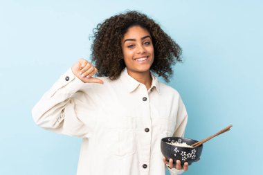 Genç Afrikalı Amerikalı bir kadın mavi arka planda kendini beğenmiş ve gururlu bir şekilde yemek çubuklarıyla bir kase erişte tutarken...