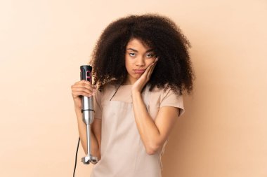 Genç Afrikalı Amerikalı kadın bej arka planda izole edilmiş el blenderi kullanıyor mutsuz ve hayal kırıklığına uğramış.