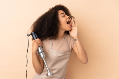 El blenderi kullanan Afrikalı genç bir kadın bej arka planda ağzı açık bağırıyor.