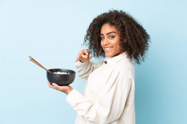 Genç Afrikalı Amerikalı bir kadın mavi arka planda izole edilmiş ve elinde çubuklarla bir kase erişte tutarken onu işaret ediyor.