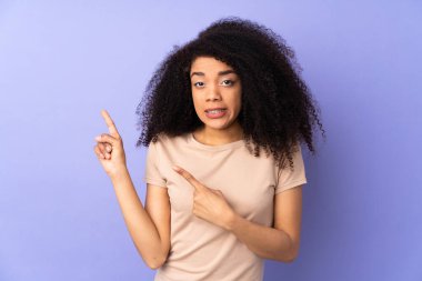 Genç Afro-Amerikalı kadın, mor arka planda izole edilmiş korkmuş ve kenarı işaret ediyor.