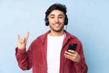 Arap yakışıklı adam mavi arka planda izole edilmiş, hareketli bir rock hareketi yaparak müzik dinliyor.