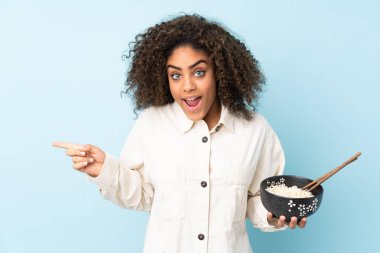 Mavi arka planda izole edilmiş genç Afrikalı Amerikalı kadın elinde çubuklarla bir kase erişte tutarken şaşırmış ve parmağıyla kenarı işaret ediyor.