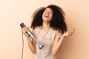 Genç Afrikalı Amerikalı kadın el blenderi kullanıyor. Bej arka planda izole edilmiş, gülerek.