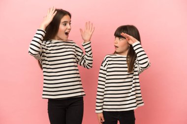 Küçük kız kardeşler pembe arka planda izole edilmiş, şaşırmış ve şok olmuş yüz ifadeleriyle.