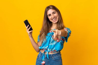 İzole edilmiş sarı arka plan üzerinde cep telefonu kullanan genç bir kadın kendinden emin bir ifadeyle seni işaret ediyor.