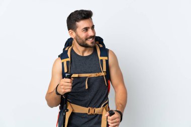 Beyaz arka planda izole edilmiş büyük bir sırt çantası ve yürüyüş direkleri olan genç dağcı yan tarafa bakıyor.