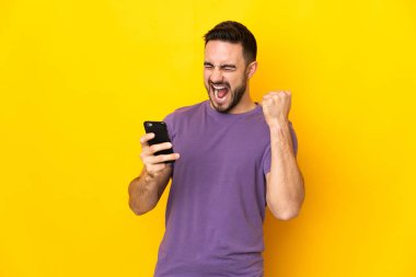 Sarı arka planda izole edilmiş, telefon zafer pozisyonunda genç beyaz yakışıklı adam.