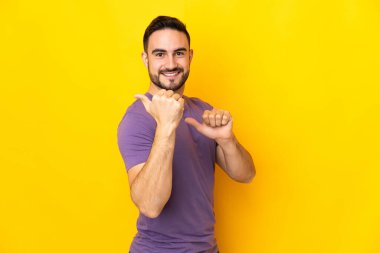 Sarı arka planda izole edilmiş beyaz, yakışıklı genç bir adam bir ürün sunmak için kenarı işaret ediyor.