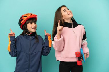 Küçük kız kardeşler bisiklet çalışıyor ve kaykaycı mavi arka planda izole edilmiş işaret parmağıyla işaret ederek harika bir fikir.