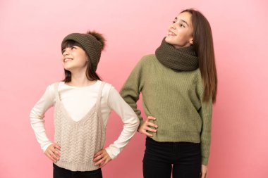 Pembe arka planda izole edilmiş, kolları kalçasında poz veren ve gülen küçük kız kardeşler.