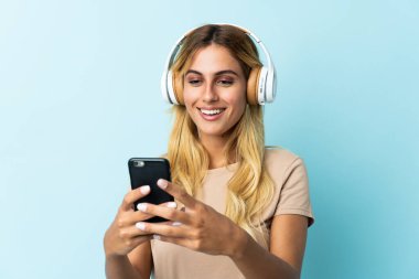 Genç sarışın Uruguaylı, mavi arka planda izole edilmiş müzik dinliyor ve cep telefonu arıyor.