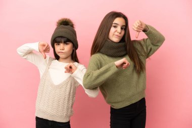 Pembe arka planda izole edilmiş kışlık giysiler giyen küçük kız kardeşler baş parmağını iki eliyle aşağı gösteriyor.