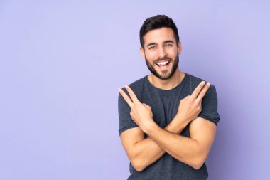 Beyaz tenli yakışıklı adam gülümsüyor ve izole edilmiş mor arka planda zafer işareti gösteriyor.