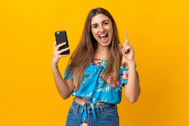 İzole edilmiş sarı arka plan üzerinde cep telefonu kullanan genç bir kadın harika bir fikre işaret ediyor.
