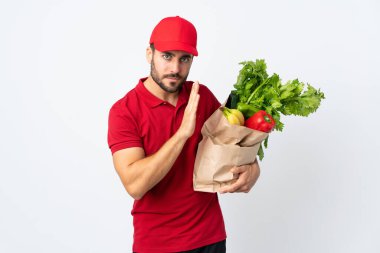 Sakallı genç adam elinde sebze dolu bir çanta beyaz arka planda izole edilmiş bir şekilde bir şeyler fısıldıyor.