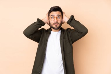 Beyaz tenli yakışıklı adam bej arka planda izole edilmiş hayal kırıklığına uğramış ve kulaklarını kapatmış.