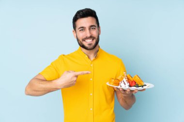 Elinde waffle tutan beyaz adam mavi arka planda izole edilmiş ve onu işaret ediyor.