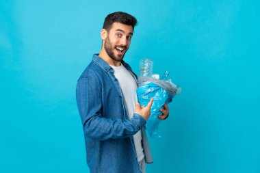 Mavi arka planda izole edilmiş plastik şişelerle dolu bir çanta taşıyan genç beyaz bir adam.