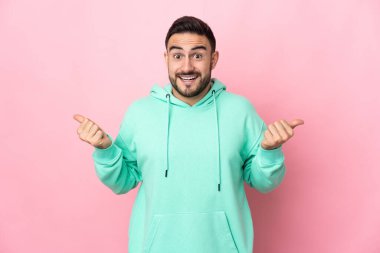 Pembe arka planda izole edilmiş beyaz, yakışıklı bir genç. Baş parmakları havada ve gülümsüyor.