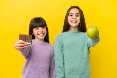 Sarı arka planda izole edilmiş küçük kız kardeşler bir elinde çikolata tableti, diğerinde elma alıyorlar.
